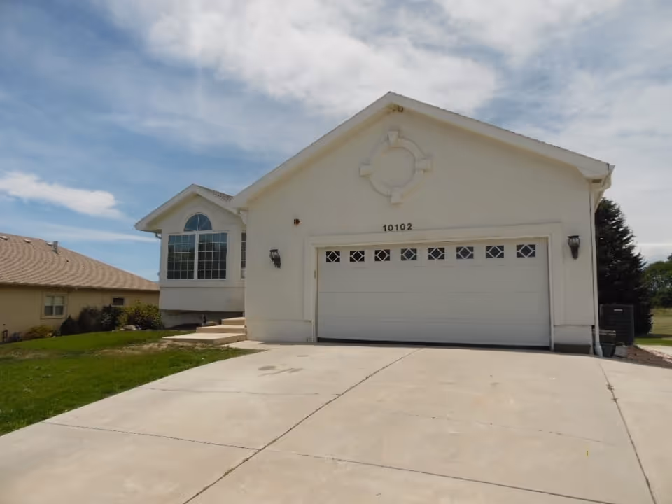 Exterior view of a single-story residential building with a large garage door, a concrete driveway, and a well-maintained lawn under a partly cloudy sky.