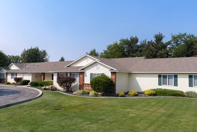 Single-story building with beige siding and a brown shingled roof, surrounded by green grass, shrubs, and trees under a clear sky.