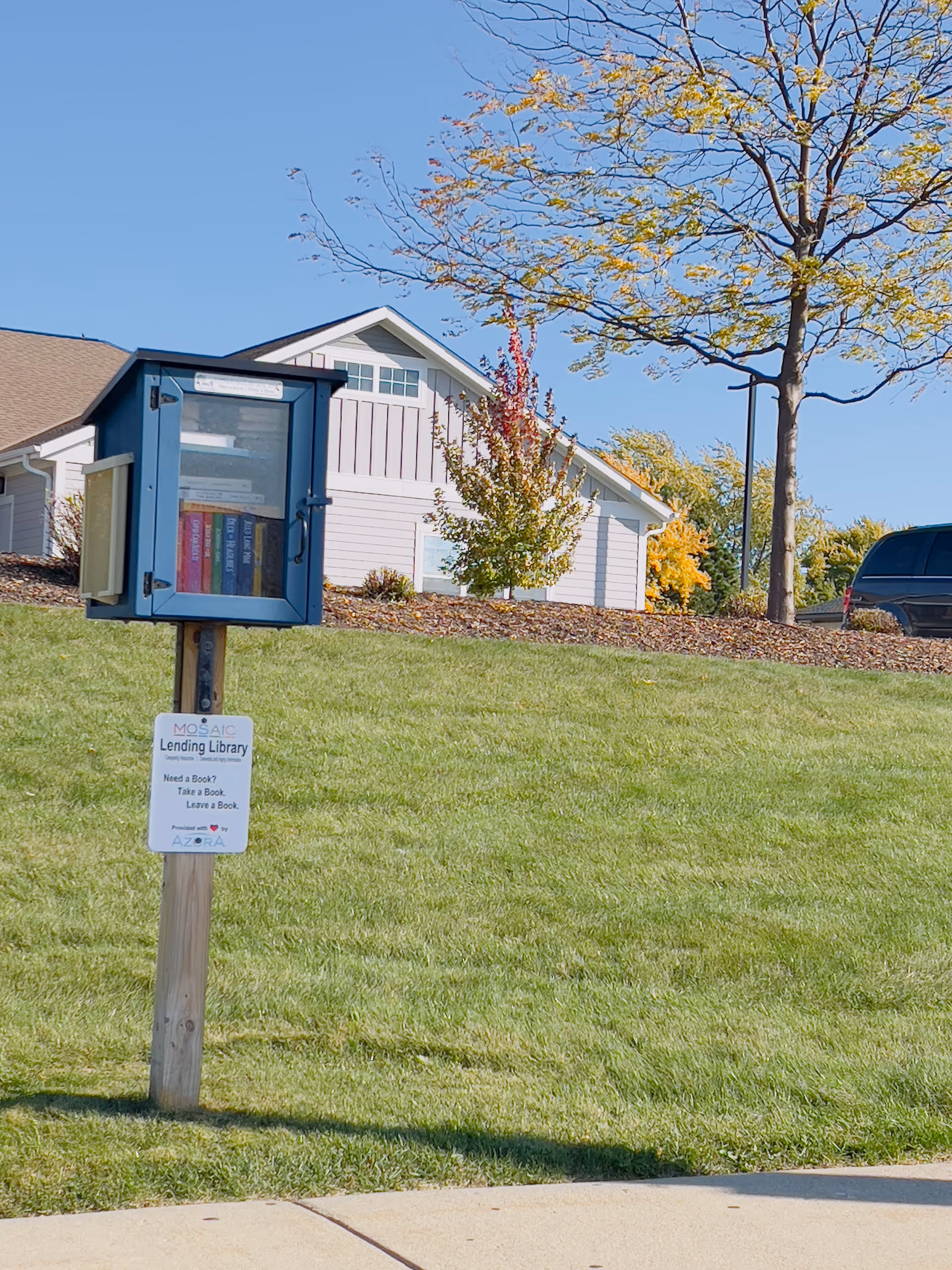 A small blue free lending library box on a post sits on a grassy lawn in front of a white building with trees and a clear blue sky.