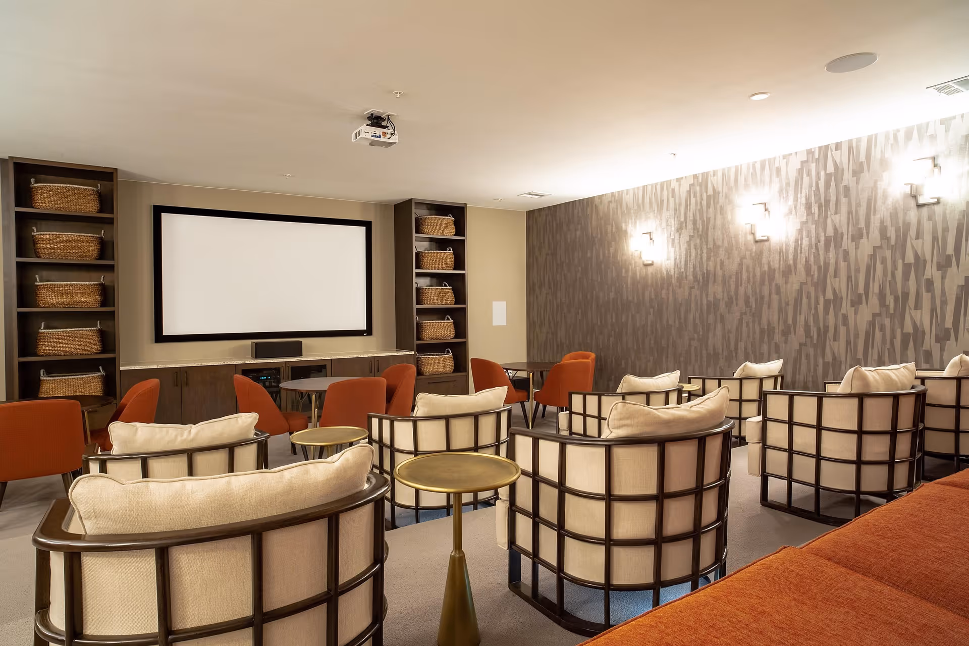 A cozy screening room with upholstered armchairs and small tables facing a large projection screen and shelving with wicker baskets.