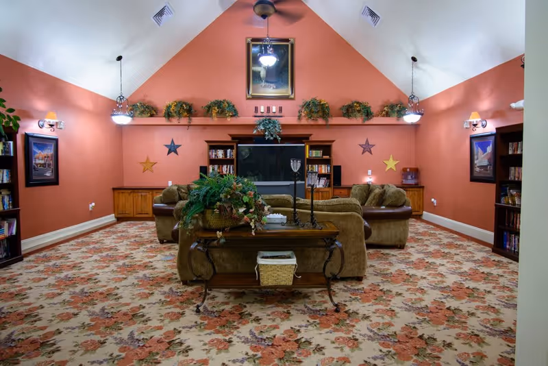 A cozy living room with floral carpet, two brown sofas facing a large flat-screen TV mounted on a coral-colored wall. The room has a vaulted ceiling with a ceiling fan and hanging light fixtures. Decorative plants and framed pictures adorn the walls and shelves.