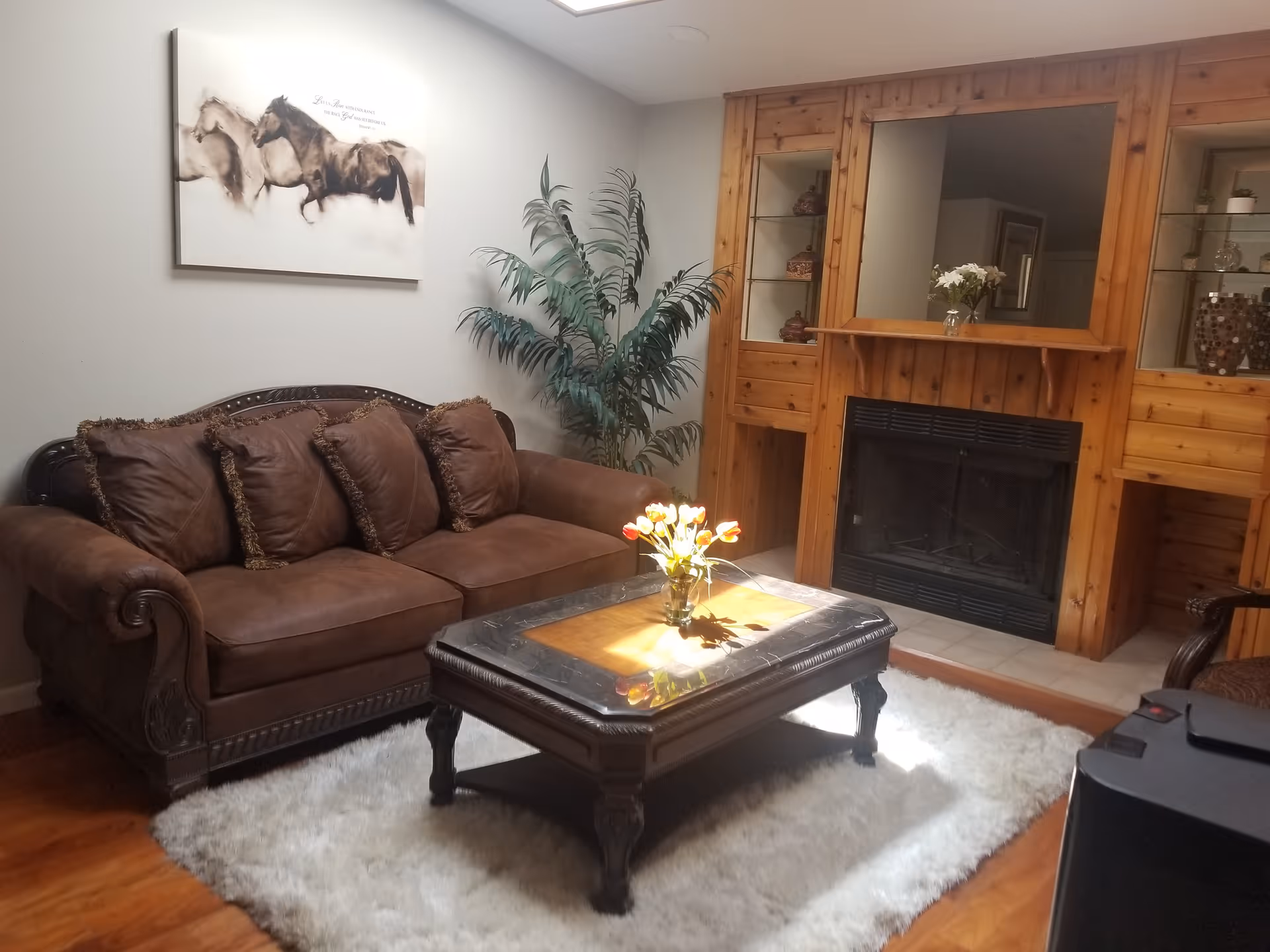 A cozy living room with a brown leather sofa adorned with four matching cushions, a wooden coffee table with a glass top holding a vase of tulips, a white fluffy rug underneath, a large green potted plant in the corner, and a wooden fireplace with built-in shelves on either side displaying decorative items. A painting of horses hangs on the wall above the sofa.