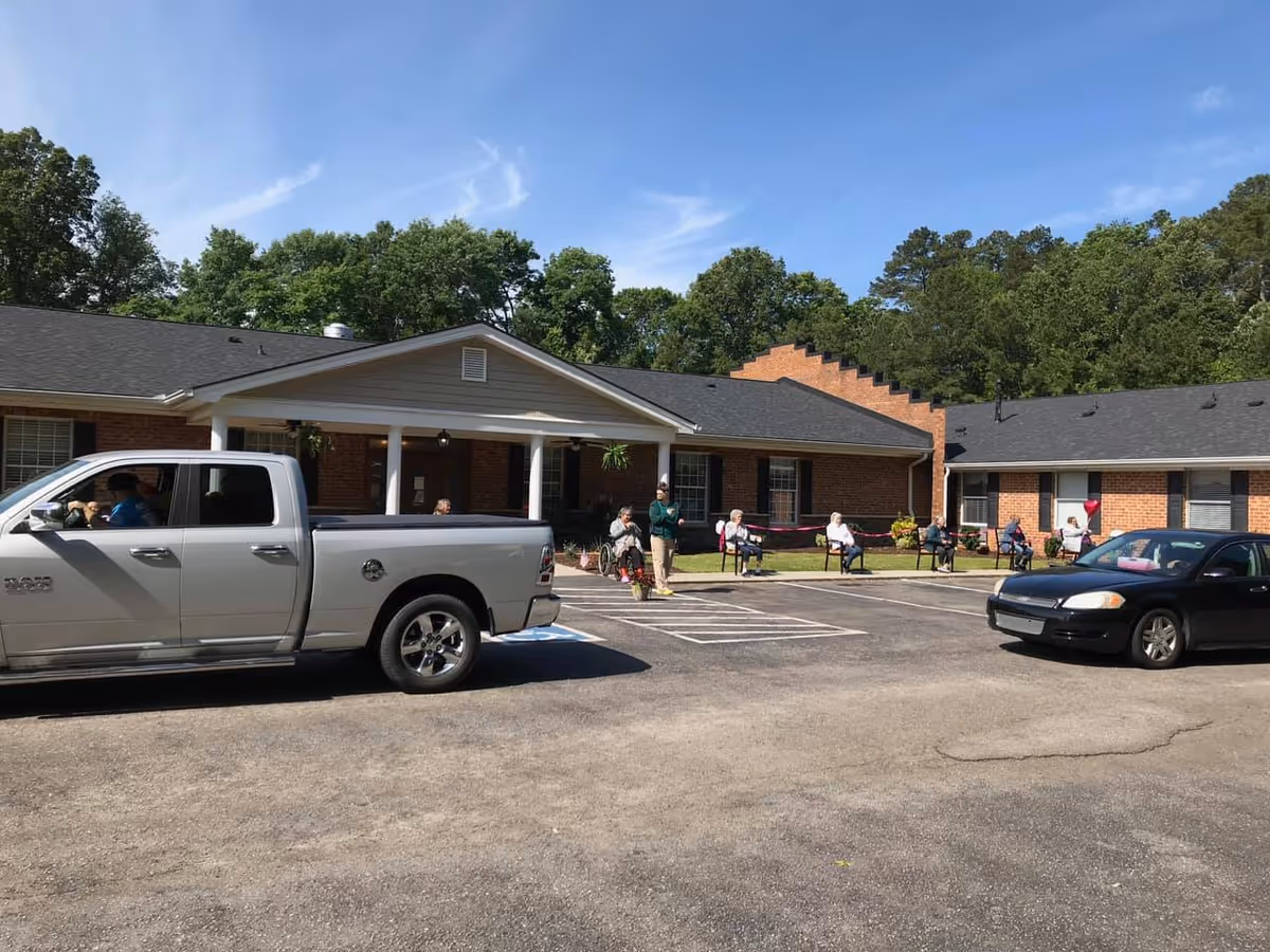 Exterior view of Lenoir Assisted Living facility showing a single-story brick building with a covered entrance. Several elderly people are seated outside on chairs spaced apart on the lawn near the building. Two vehicles, a silver pickup truck and a black sedan, are parked in front of the building. Trees and a clear blue sky are visible in the background.