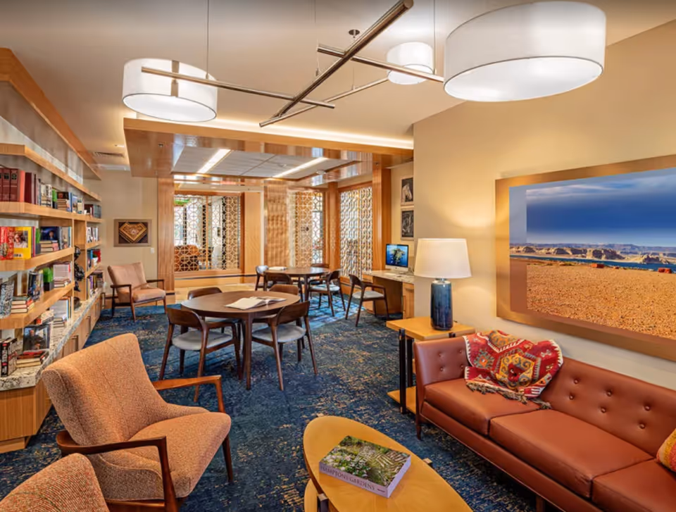 A cozy and well-lit common area in a senior living facility featuring a brown leather sofa with colorful pillows, a wooden coffee table with a book, two upholstered armchairs, round wooden tables with chairs, a bookshelf filled with books, a desk with a computer, and decorative wall art. The room has a blue patterned carpet and modern ceiling lights.