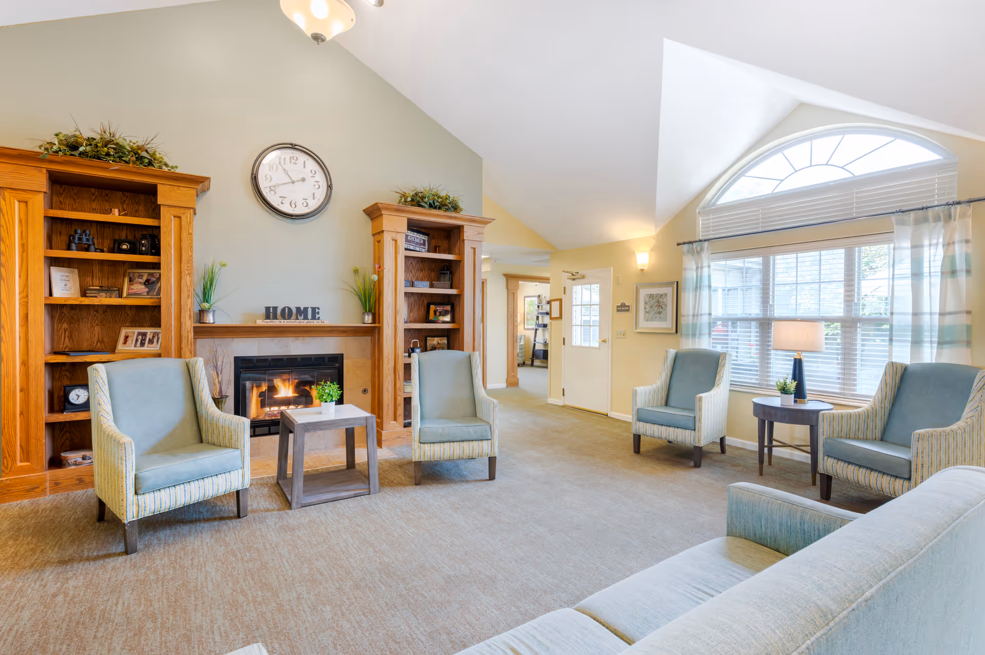 A bright and cozy living room area with a fireplace in the center flanked by two wooden bookshelves. Four upholstered armchairs and a sofa are arranged around two small tables. A large arched window with blinds and curtains lets in natural light. The walls are painted in soft neutral tones, and there is a clock above the fireplace along with decorative plants and framed photos.