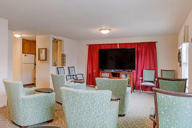 A common area in a senior living facility with several green patterned armchairs arranged facing a large flat-screen TV mounted on a wooden stand. The room has beige walls, a carpeted floor, and a window with red curtains behind the TV. To the left, a small kitchen area with a refrigerator is partially visible.