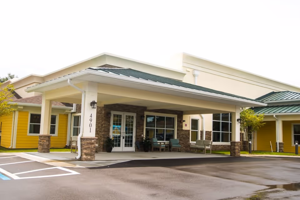 Covered front entrance of a yellow-and-stone senior living building with seating under a porte-cochère and the address number 4901 on a column.