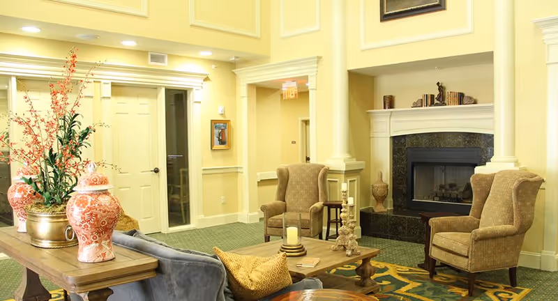 A cozy living room area with two upholstered armchairs facing a fireplace with a black marble surround. A wooden coffee table with candles and books sits in front of the chairs. A sofa with yellow patterned pillows is partially visible. Decorative vases and a plant are on a wooden console table behind the sofa. The walls are painted light yellow, and the room has green carpet and white trim.