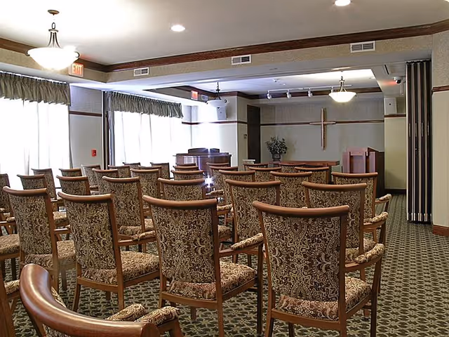 A room set up with multiple rows of patterned upholstered chairs facing a small stage area with a wooden cross on the wall and a podium, illuminated by ceiling lights and natural light from large windows with curtains.