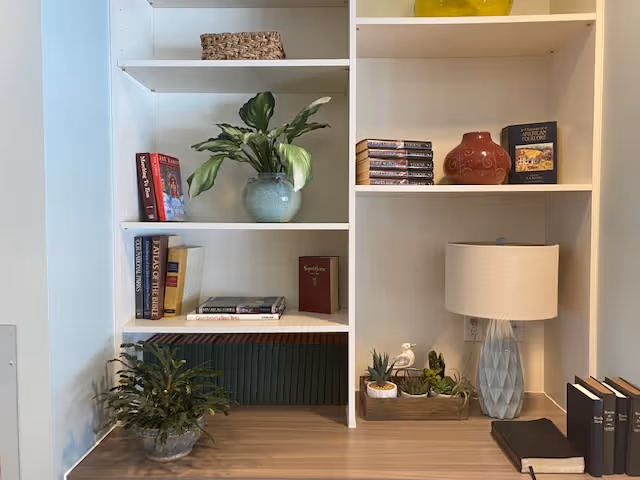 A white built-in bookshelf with various decorative items including potted plants, books, a table lamp with a white shade, and a red ceramic vase. The shelves are neatly arranged with a mix of greenery and reading materials.
