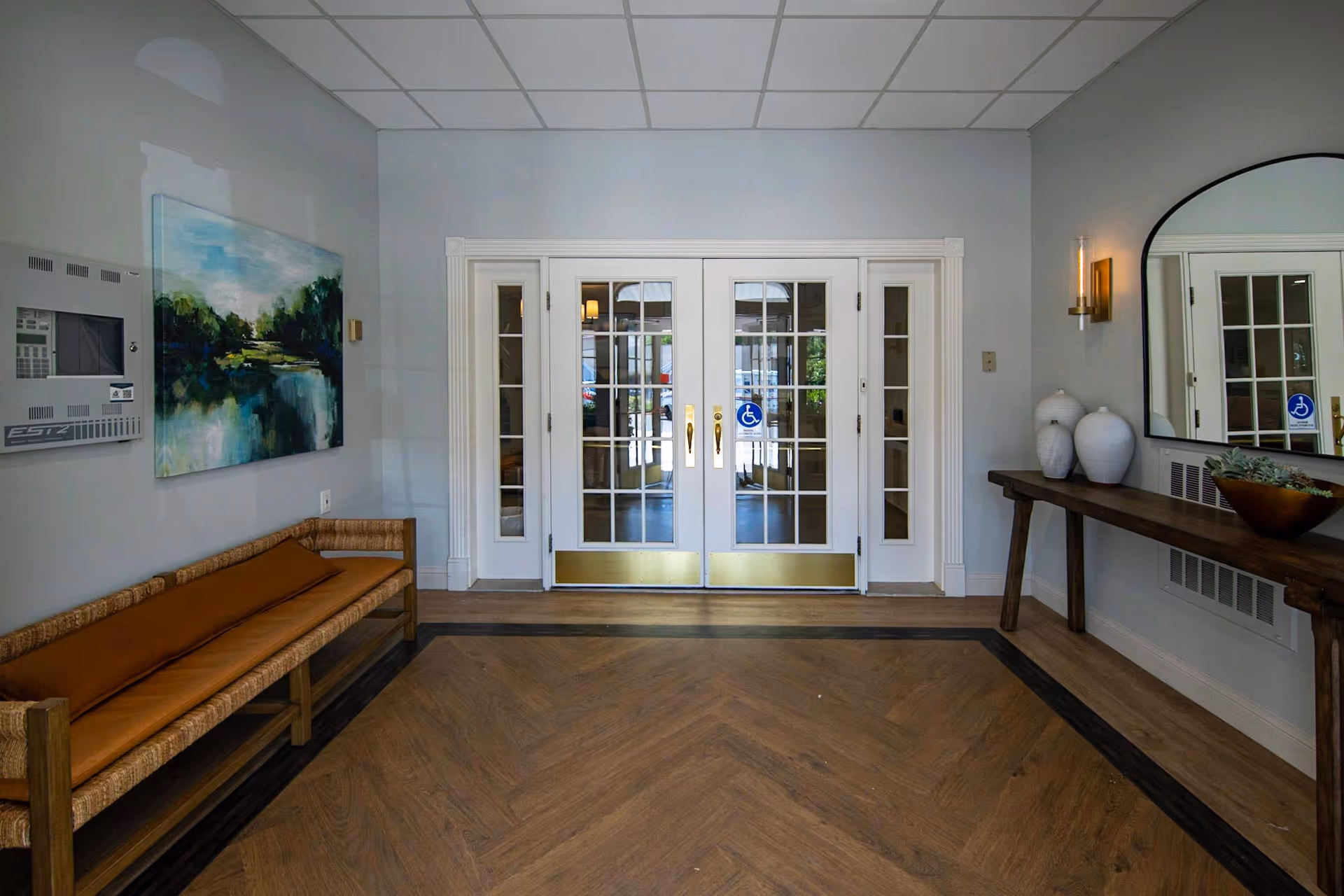 Interior view of an entrance area with double glass doors featuring gold handles and handicap accessibility stickers. To the left is a wooden bench with a brown cushion and a landscape painting above it. To the right is a wooden console table with decorative vases and a bowl of succulents, a large mirror above it, and a wall-mounted light fixture. The floor has a wood pattern with a dark border, and the ceiling has a grid of white tiles.