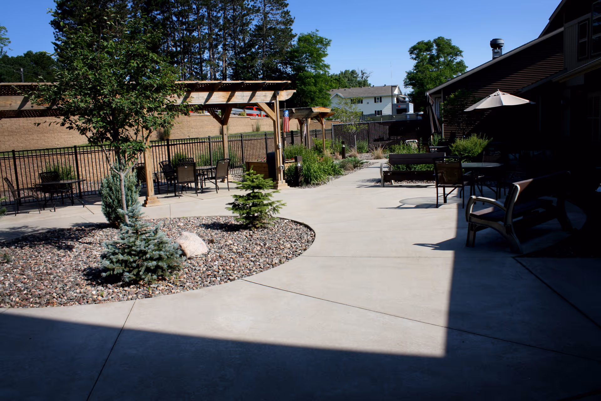 Outdoor patio area with concrete walkways, small landscaped garden beds with rocks and small trees, several seating areas with chairs and tables, wooden pergolas, and a building with an umbrella-covered table on the right side under clear blue sky.