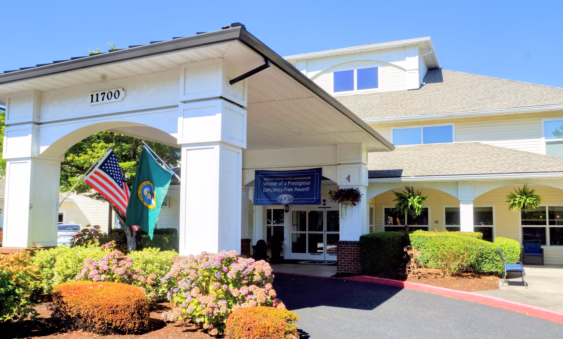 Front entrance and porte-cochère of a senior living building marked 11700, with American and state flags, hanging plants, and landscaped flower beds.