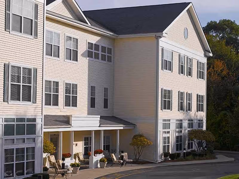 Exterior view of a multi-story senior living facility building with beige siding, multiple windows, a covered entrance with seating, and some landscaping including small trees and bushes.