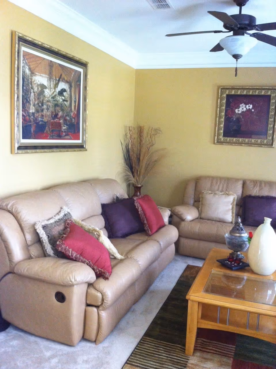 A living room with two beige leather sofas adorned with red and purple decorative pillows. There is a wooden coffee table with decorative items including a white vase and a small ornate container. The walls are painted yellow and decorated with two framed paintings. A ceiling fan with a light fixture is mounted on the ceiling.