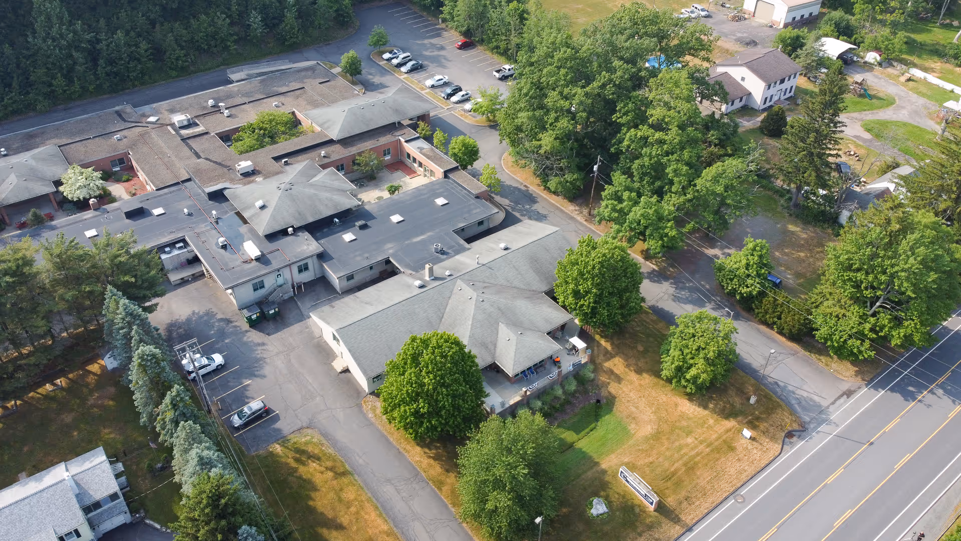 Aerial view of Mountain Top Rehabilitation and Healthcare Center showing a large building complex surrounded by trees, parking lots with several cars, and adjacent roads. The facility is set in a green area with well-maintained lawns and some smaller buildings nearby.