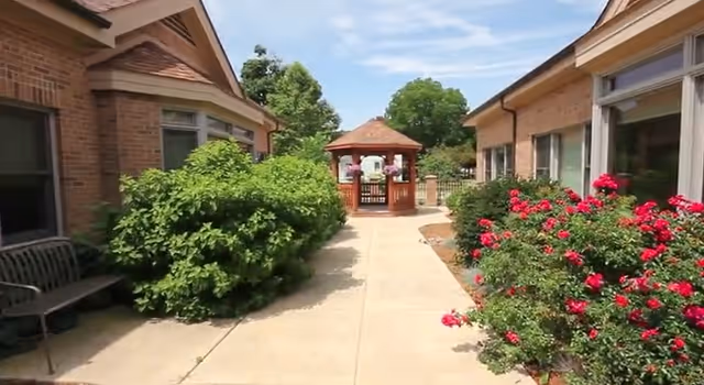 Outdoor walkway between two brick buildings with green bushes on the left and red flowering bushes on the right, leading to a wooden gazebo under a partly cloudy sky.