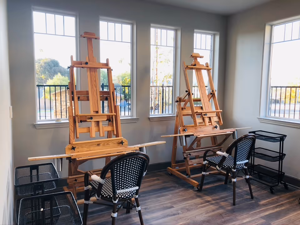 A bright room with three tall windows letting in natural light, containing two wooden easels and two black and white woven chairs. There are also two black metal rolling carts with mesh baskets on either side of the easels. The floor is wood and the walls are painted light gray.