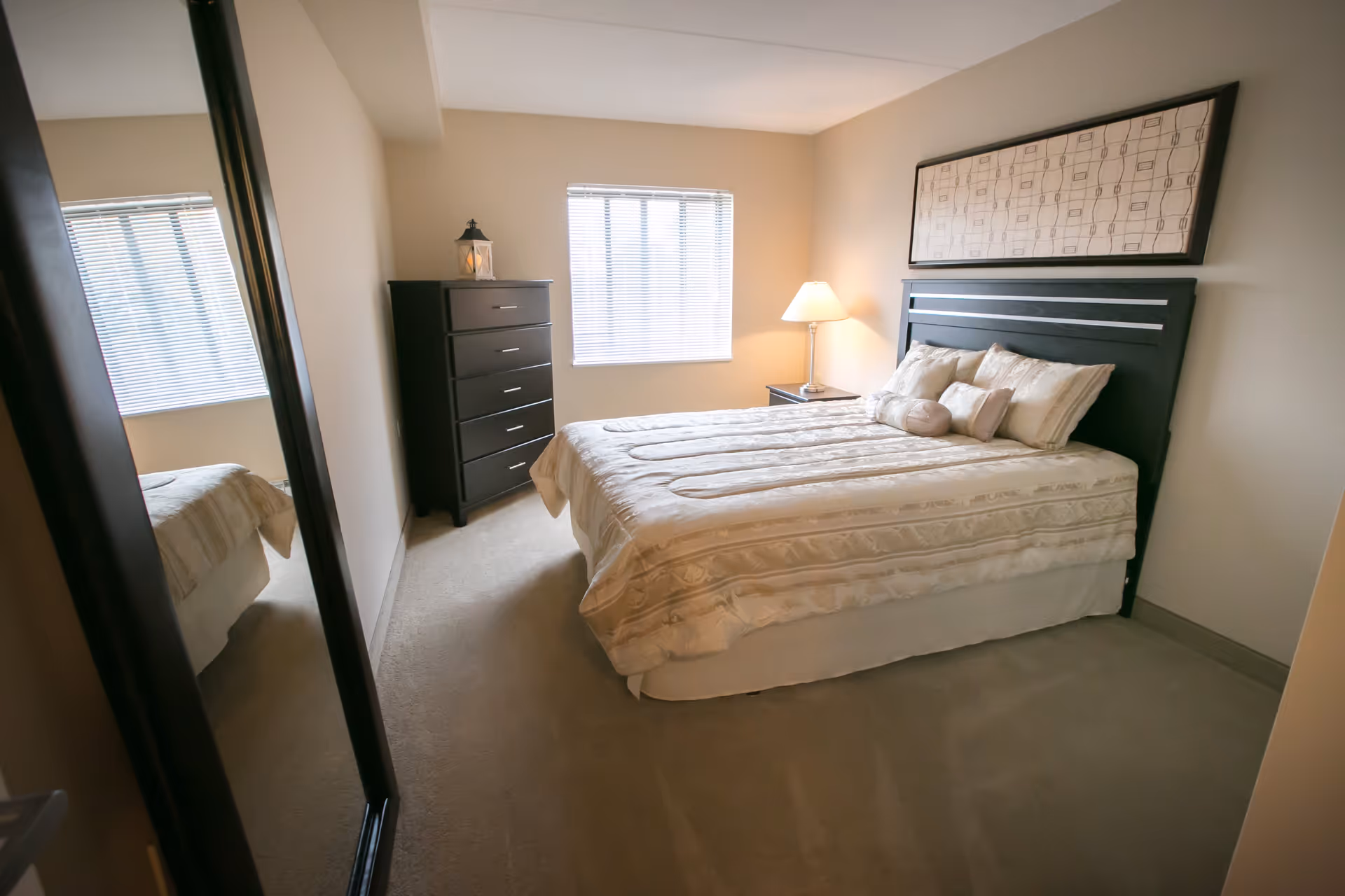 A neatly made bed with beige and cream bedding in a bedroom. There is a black headboard with a framed decorative piece above it. To the right of the bed is a nightstand with a lamp. Across from the bed is a tall black dresser with a decorative lantern on top. A window with blinds lets in natural light, and a large mirror is visible on the left wall.