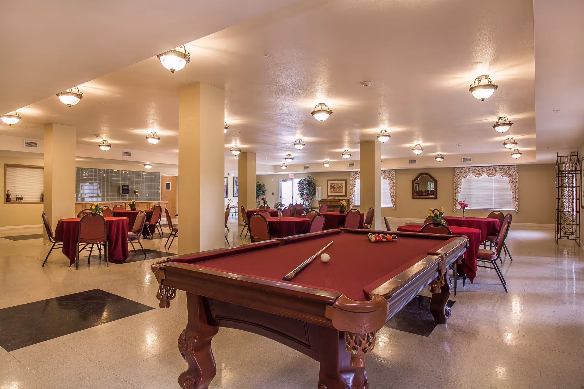 A spacious senior living common area with a pool table in the foreground and several round tables covered with red tablecloths and chairs in the background. The room is well-lit with multiple ceiling lights, beige walls, and windows with decorative curtains. There are plants and framed pictures on the walls, creating a welcoming atmosphere.