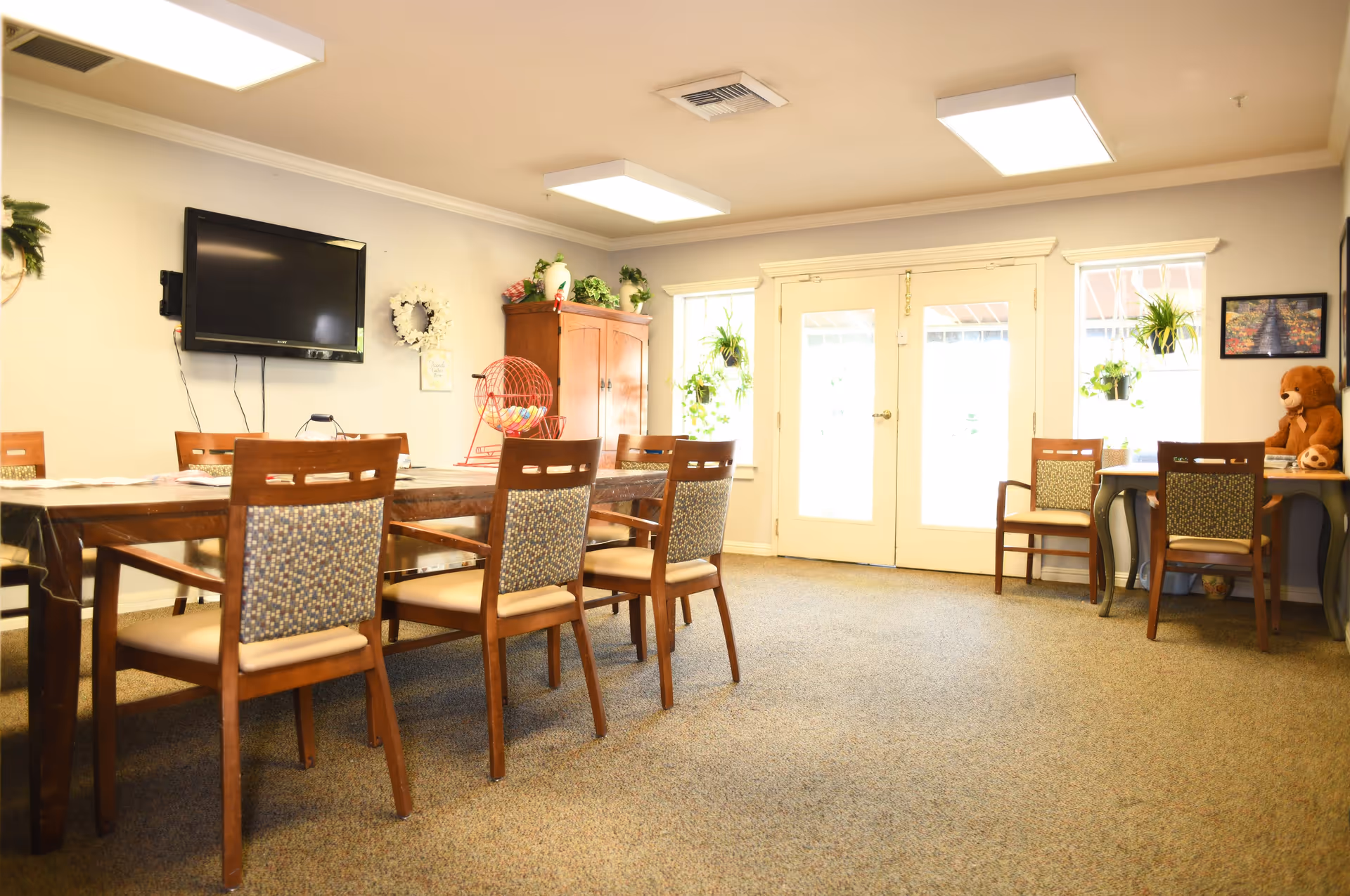 A bright room with a long wooden table surrounded by chairs with patterned cushions. On the table is a bingo cage with colorful balls inside. The room has a wall-mounted flat screen TV, a wooden cabinet with plants and decorations on top, and double glass doors letting in natural light. There is also a small table with chairs, a large teddy bear, and hanging plants near the windows.