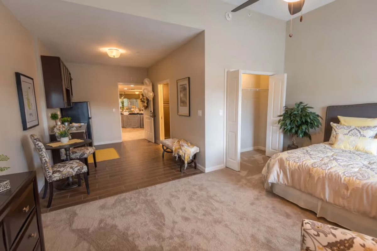 A well-lit bedroom with a bed adorned with patterned yellow and white bedding, a green potted plant beside the bed, an open closet with double doors, and a small seating area with two floral-patterned chairs and a round table. The room has a mix of carpet and wood flooring, and a ceiling fan is visible above. In the background, there is a hallway leading to another room with balloons near the doorway.