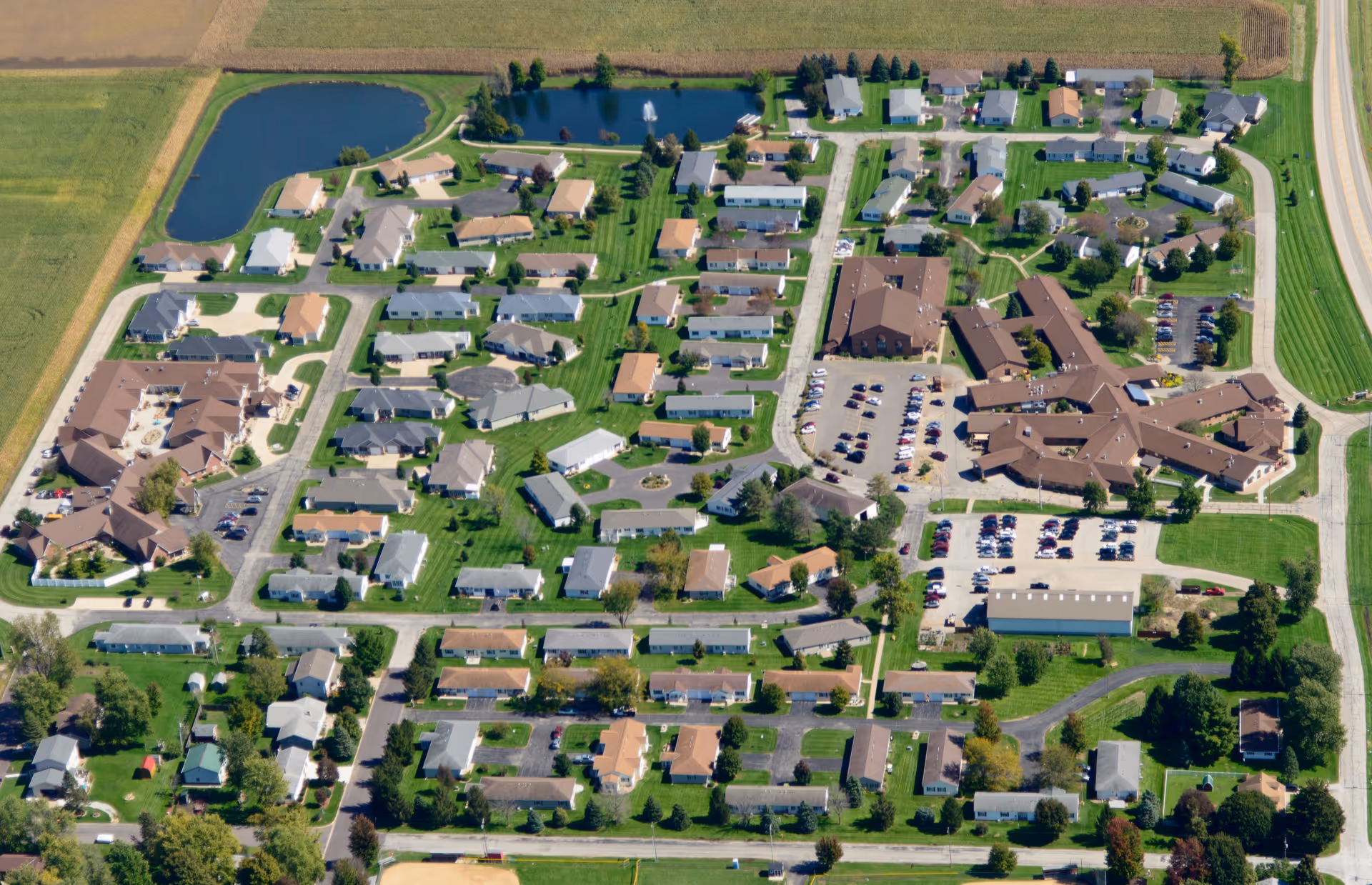 Aerial view of Snyder Village senior living facility showing multiple residential buildings, parking lots, green lawns, trees, and a pond with a fountain, surrounded by farmland.