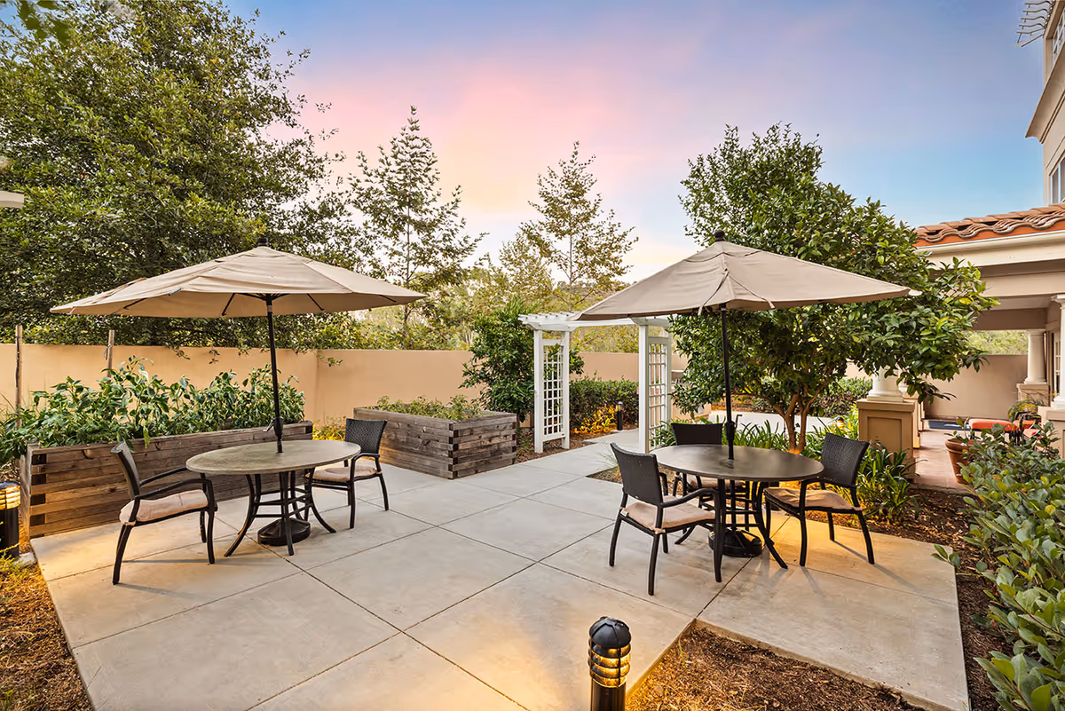 Outdoor patio area with two round tables, each shaded by a large beige umbrella and surrounded by four chairs. The patio is paved with concrete and bordered by greenery, including trees, bushes, and raised wooden garden beds. A white trellis archway is visible in the background under a colorful sunset sky.