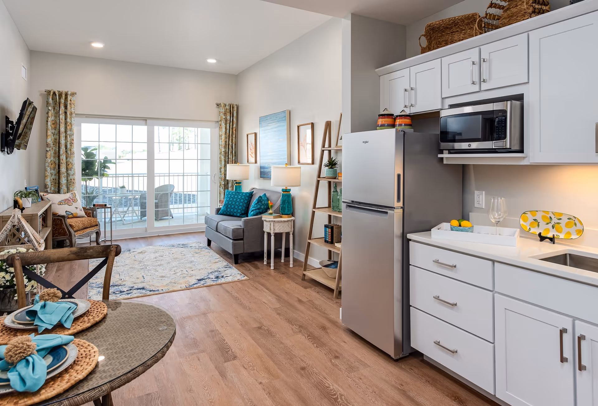 Bright and modern open-concept living space featuring a small kitchen with white cabinets, a stainless steel refrigerator, and a microwave. Adjacent to the kitchen is a cozy living area with a gray loveseat adorned with teal pillows, two matching table lamps, and wall art. A sliding glass door leads to an outdoor patio with seating. A round dining table with place settings is visible in the foreground.