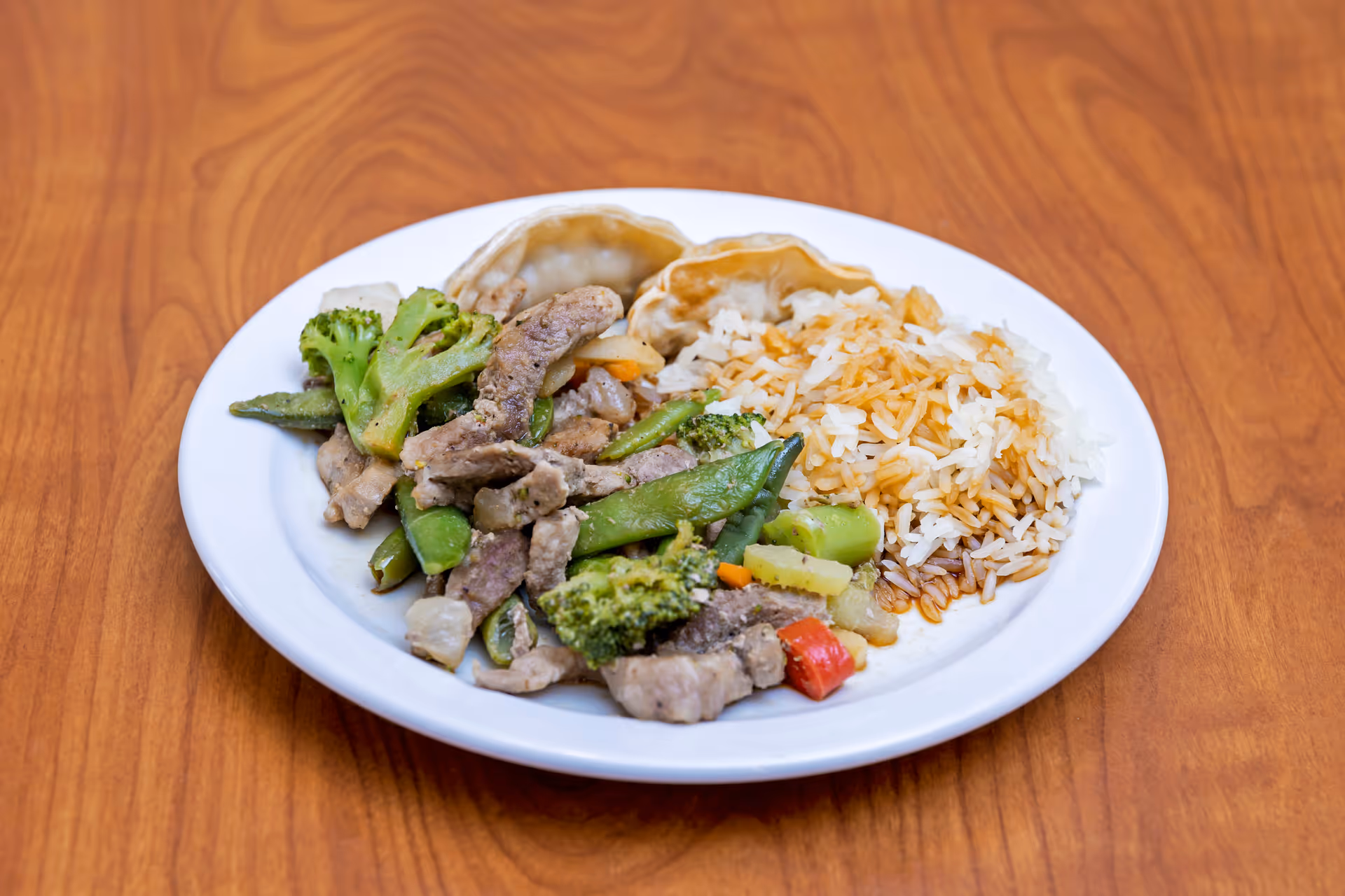 A white plate on a wooden table containing cooked white rice with soy sauce, steamed mixed vegetables including broccoli and snap peas, pieces of cooked meat, and two dumplings.