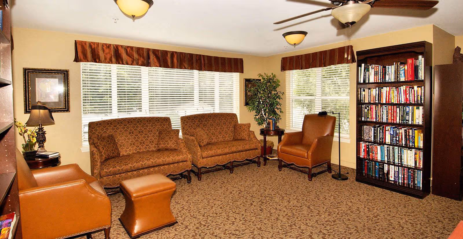 A cozy living room with two patterned sofas, two brown leather armchairs, a matching leather ottoman, a tall bookshelf filled with books, a small side table with a lamp and plant, and large windows with blinds and valances letting in natural light.