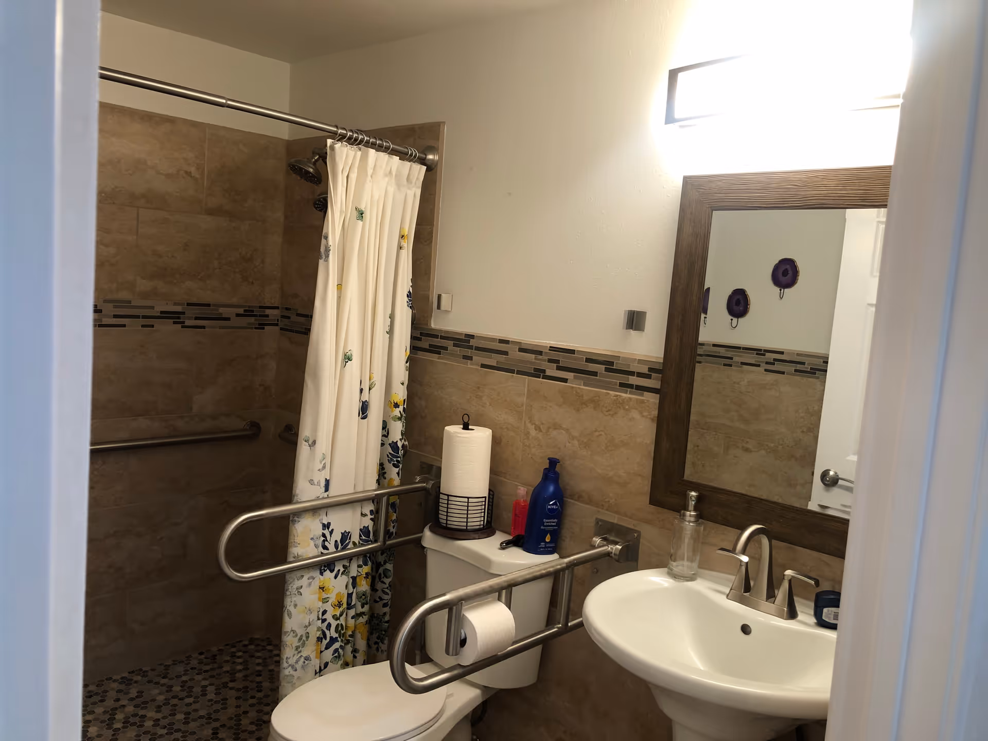 A small bathroom with beige tiled walls and floor. There is a white toilet with metal safety rails on both sides, a roll of paper towels on top of the toilet tank, and a toilet paper roll on the rail. Next to the toilet is a white pedestal sink with a silver faucet and a soap dispenser. Above the sink is a rectangular mirror with a wooden frame. To the left is a shower area with a floral shower curtain and a silver showerhead. The lighting is bright from a fixture above the mirror.