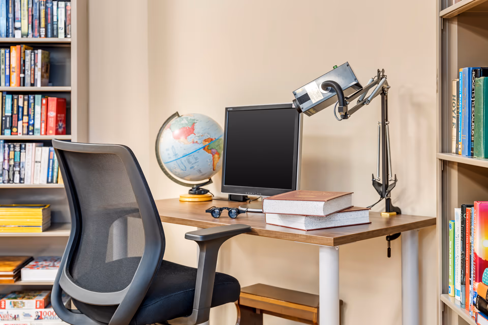 A study or office area with a black mesh office chair, a wooden desk holding a globe, a computer monitor, a desk lamp, two large books, and a pair of glasses. Bookshelves filled with books are visible on both sides of the desk.