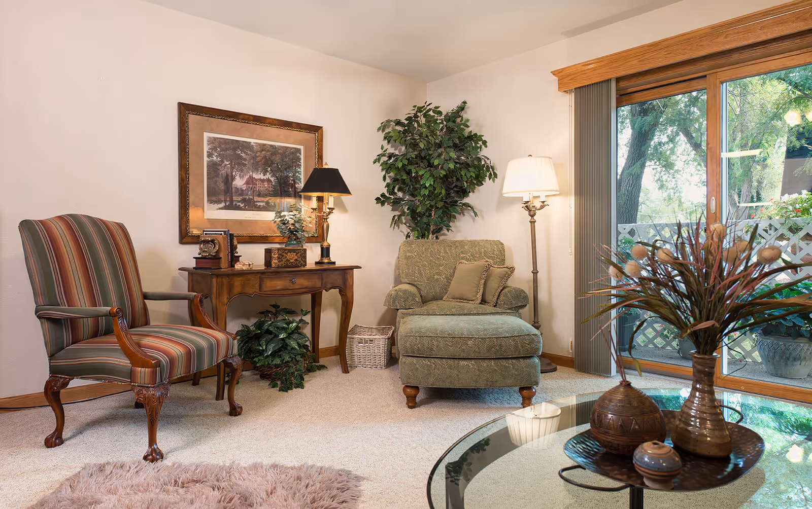 Cozy living room with an upholstered armchair and ottoman, a striped accent chair, side table with a lamp, plants, and sliding glass doors to a patio.