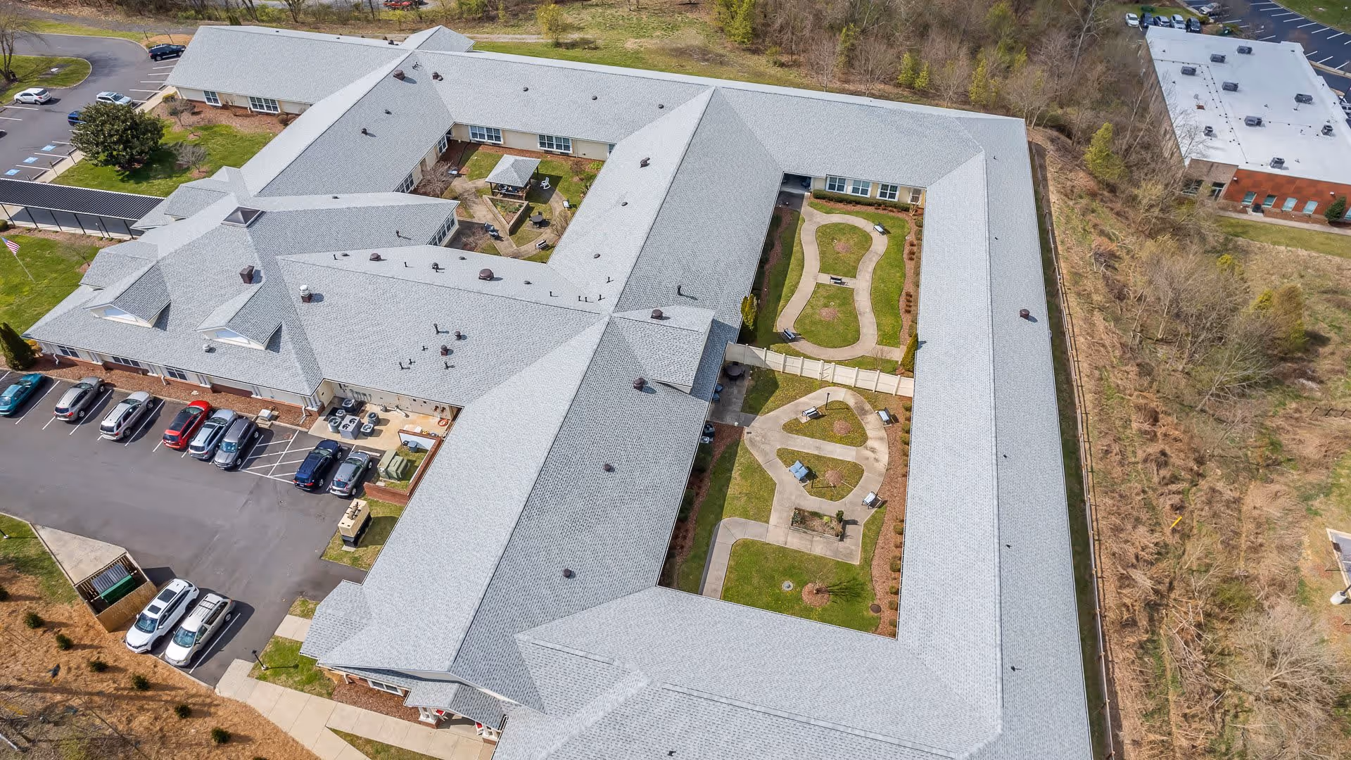 Aerial view of American House Bristol senior living facility showing a large building with a gray roof surrounding two landscaped courtyards with walking paths, benches, and greenery. There is a parking lot with several cars on one side and wooded areas surrounding the property.