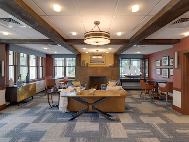 A cozy living room area with a large brown leather sofa facing a fireplace. The room features wooden ceiling beams, a modern chandelier, and large windows letting in natural light. There are decorative items on the mantel and a model sailboat on a side table. The walls are painted in warm tones with framed pictures, and there are several chairs and tables arranged around the room.