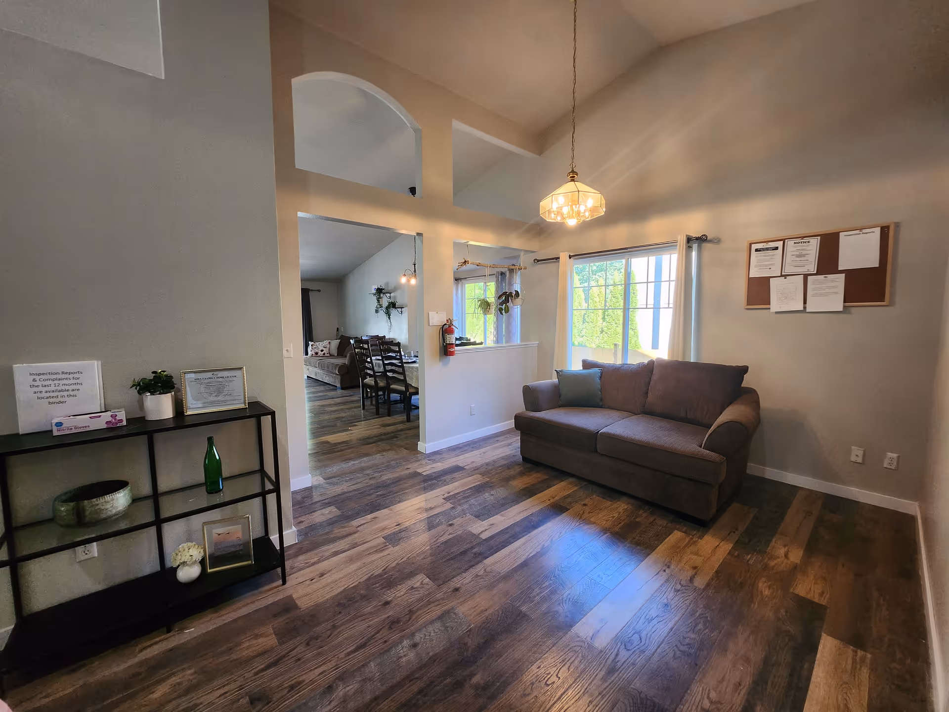 Interior view of a senior care home living area with a brown sofa, a window with white curtains, a hanging light fixture, a black shelving unit with decorative items, and a bulletin board on the wall. The room has wooden flooring and an open archway leading to a dining area with a table and chairs.