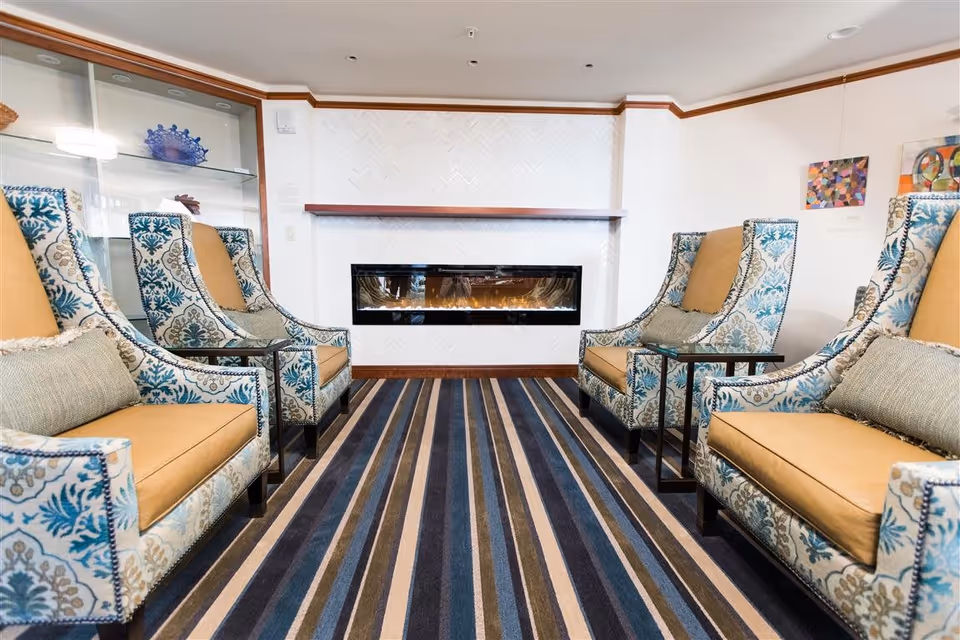 A cozy sitting area with four patterned armchairs arranged around a modern electric fireplace mounted on a white herringbone tiled wall. The room features a striped carpet in shades of blue, beige, and brown, with small side tables next to each chair and decorative art pieces on the walls.