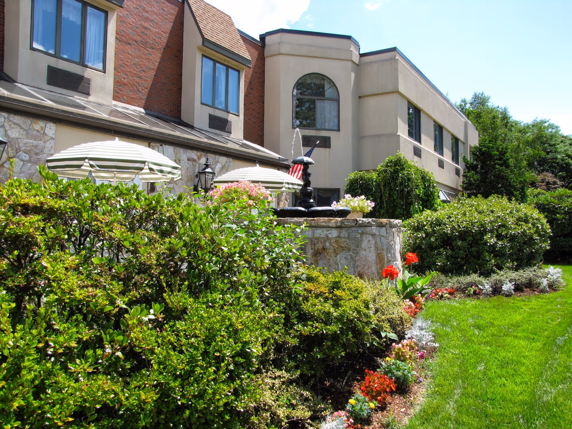 Outdoor garden area of a senior living facility with green bushes, colorful flowers, a stone fountain with water flowing, striped patio umbrellas, and a two-story building in the background under a clear blue sky.