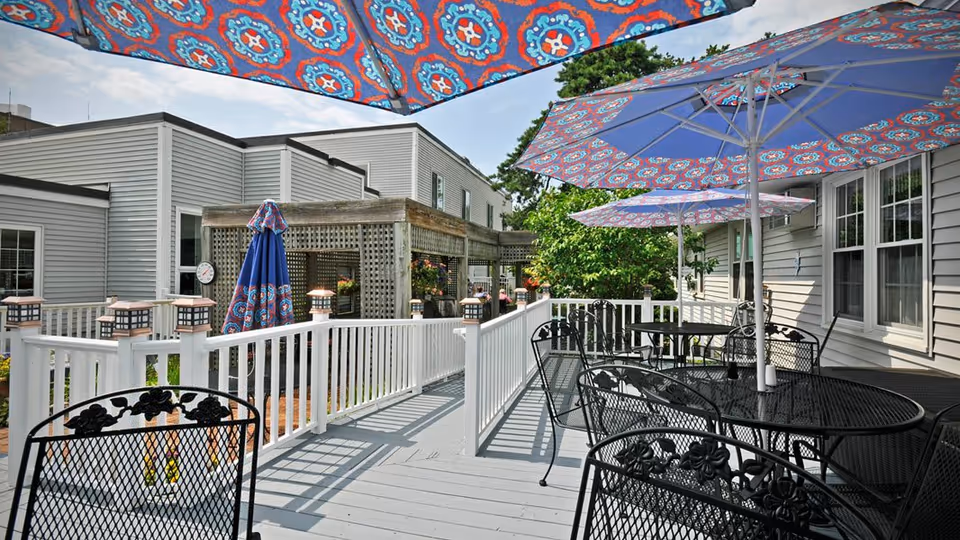 Sunlit outdoor patio deck with black metal tables and chairs and colorful patterned umbrellas beside a light-gray building.