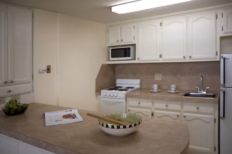Bright kitchen with white cabinets, a stove and microwave, sink, refrigerator, and a countertop island with a bowl and magazine.