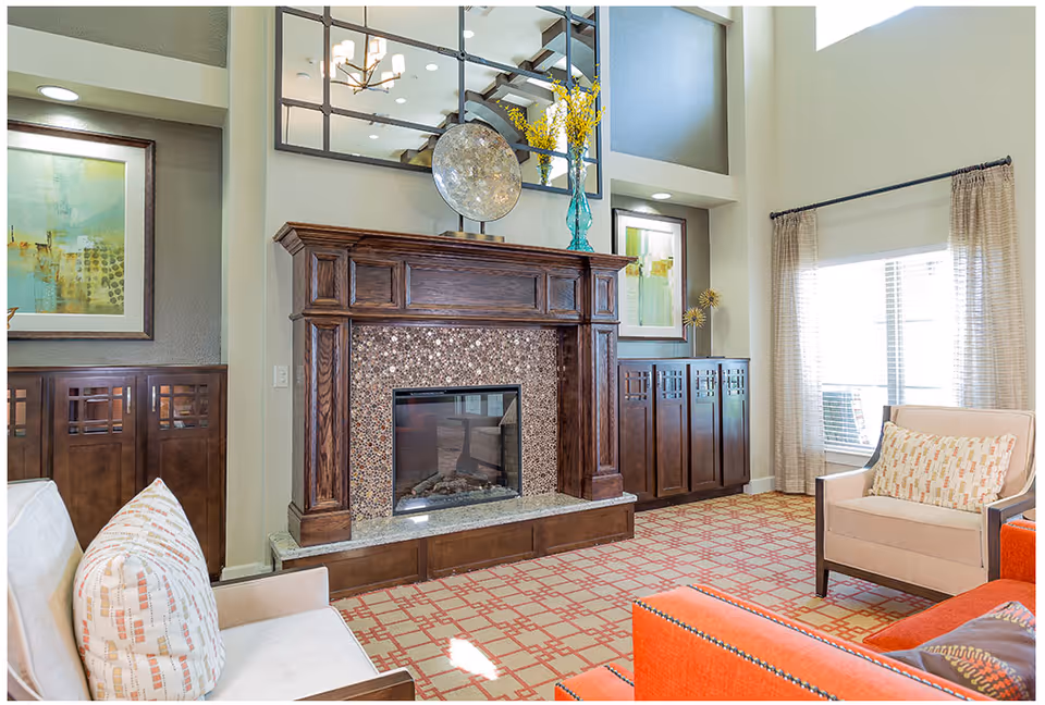 A bright living room with a large wooden fireplace featuring a mosaic tile surround. The room has patterned carpet, beige armchairs with patterned cushions, an orange sofa, and large windows with beige curtains letting in natural light. Decorative artwork and a large mirror are mounted above the fireplace.