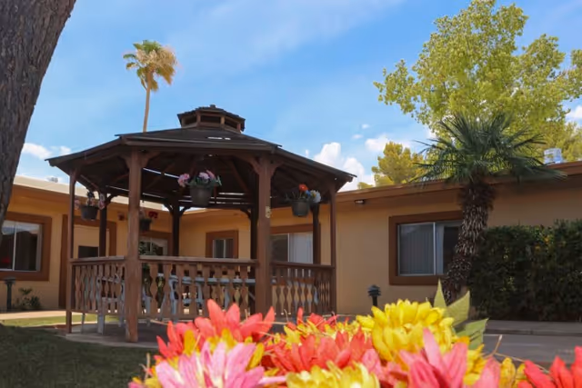 Wooden gazebo in a sunny courtyard in front of a single-story facility building with colorful flowers in the foreground.
