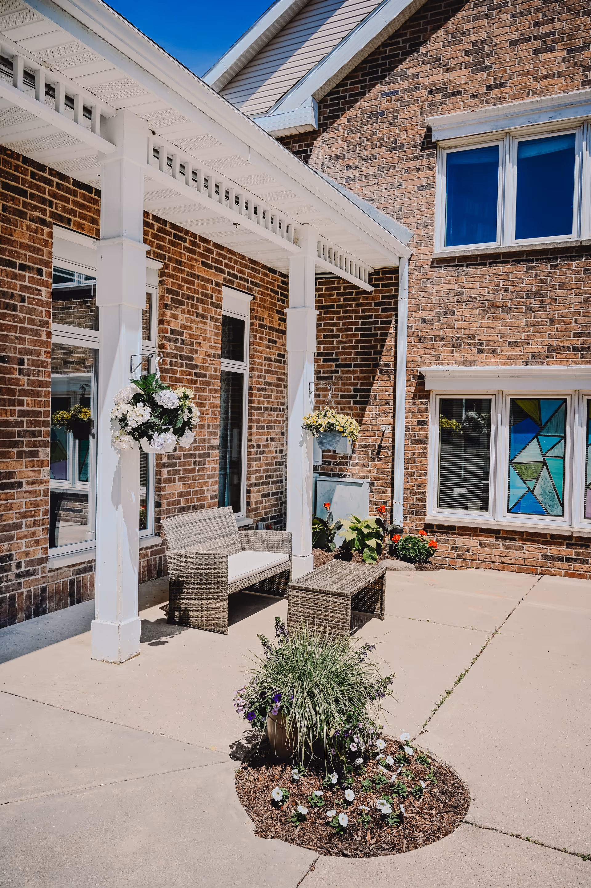 Outdoor patio area at Eden Vista Madison featuring a wicker bench with white cushions and a matching wicker coffee table under a white pergola attached to a brick building. Hanging flower baskets and a small circular flower bed with various plants and flowers are also visible.