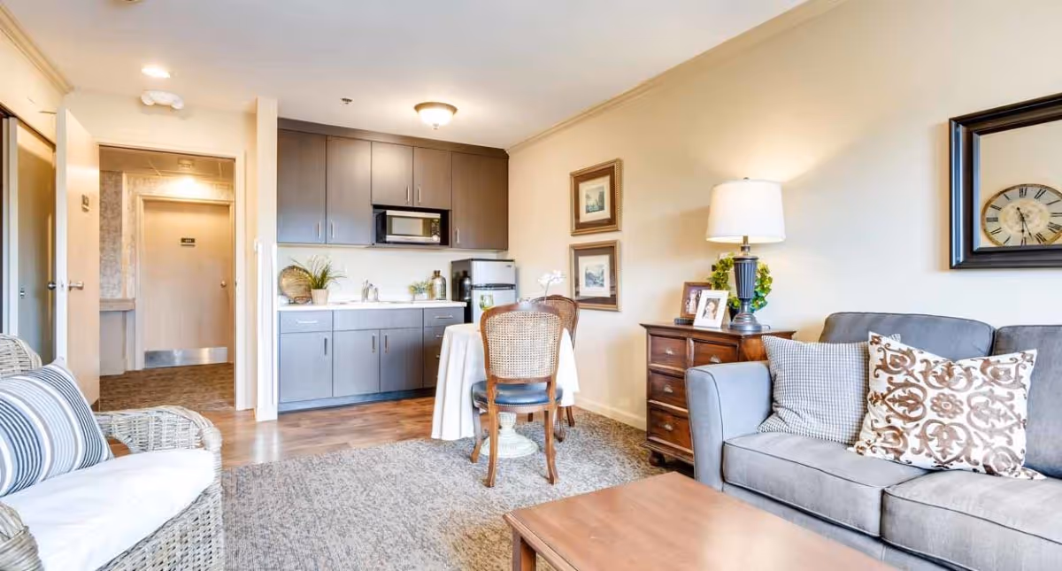 A cozy living area in an assisted living community featuring a gray sofa with patterned pillows, a wooden coffee table, a side table with a lamp and framed photos, a small dining table with two chairs, and a kitchenette with gray cabinets, a microwave, and a mini fridge. The room has light-colored walls and a carpeted floor with a wooden section near the kitchenette.