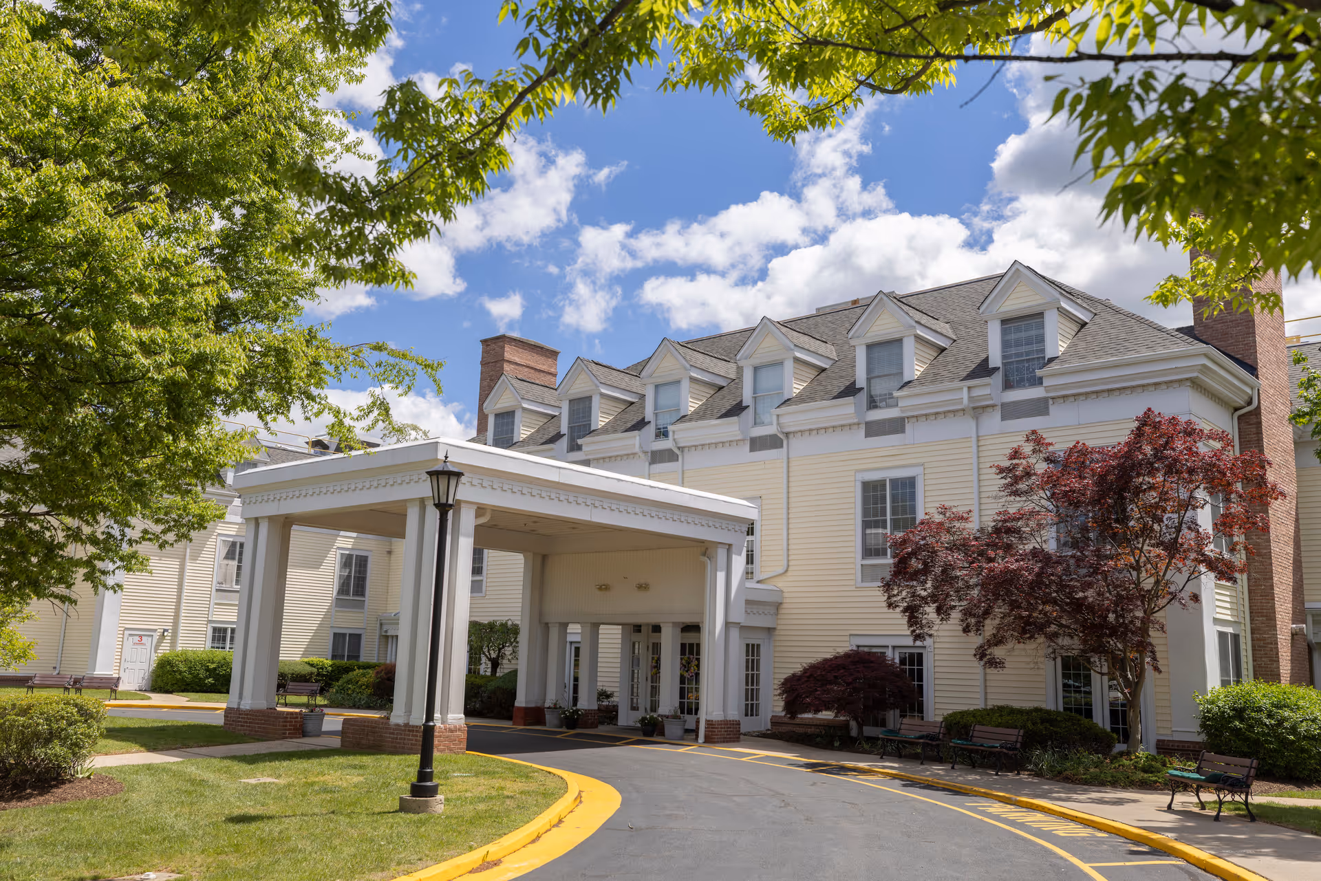 Exterior view of a senior living facility building with a covered entrance driveway. The building is light yellow with white trim and multiple dormer windows on the roof. There are green trees and shrubs around the building, and a clear blue sky with some clouds overhead.