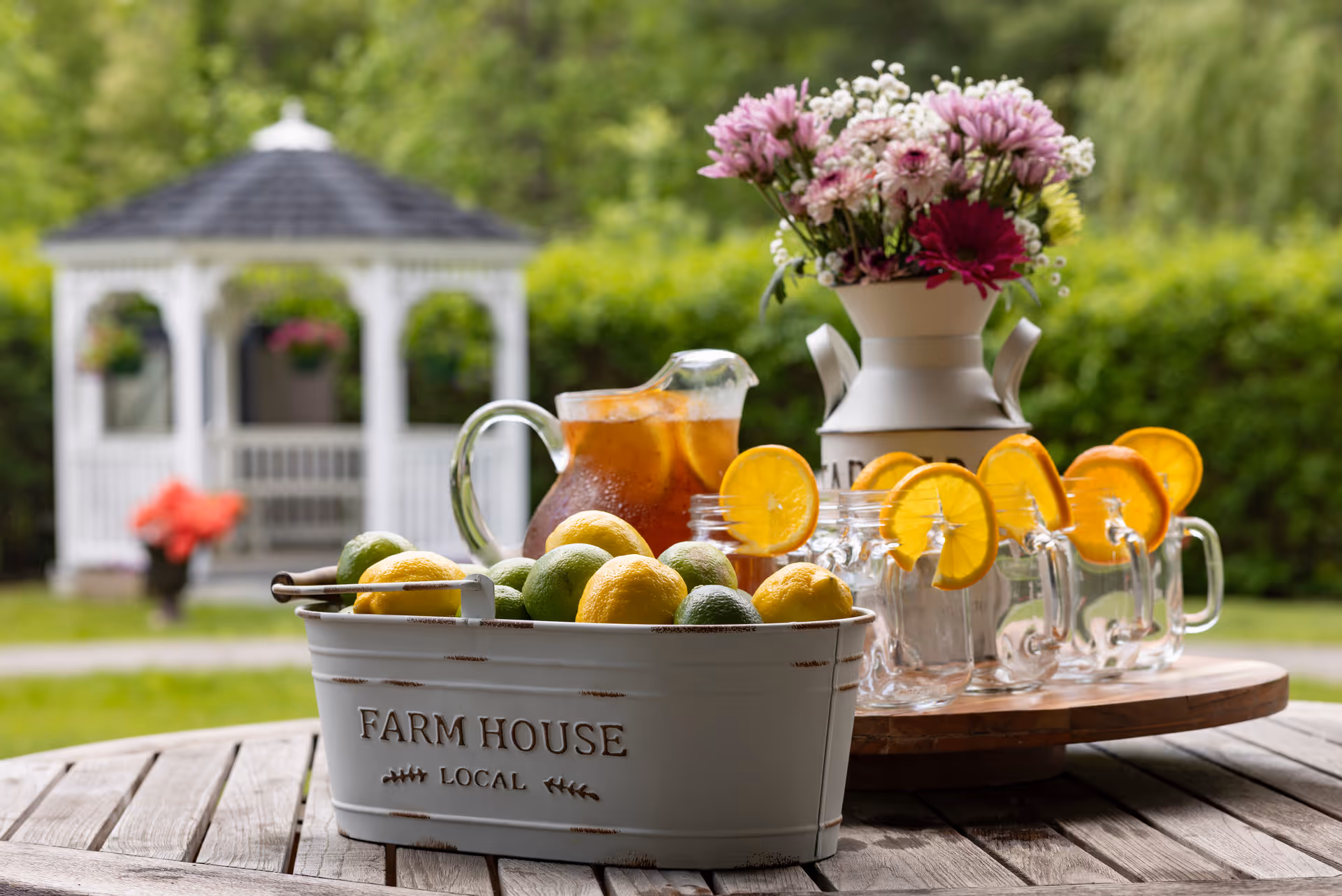 A wooden outdoor table set with a bucket of lemons and limes, a pitcher and mason jars of iced tea garnished with orange slices, a vase of flowers, and a white gazebo in the background.