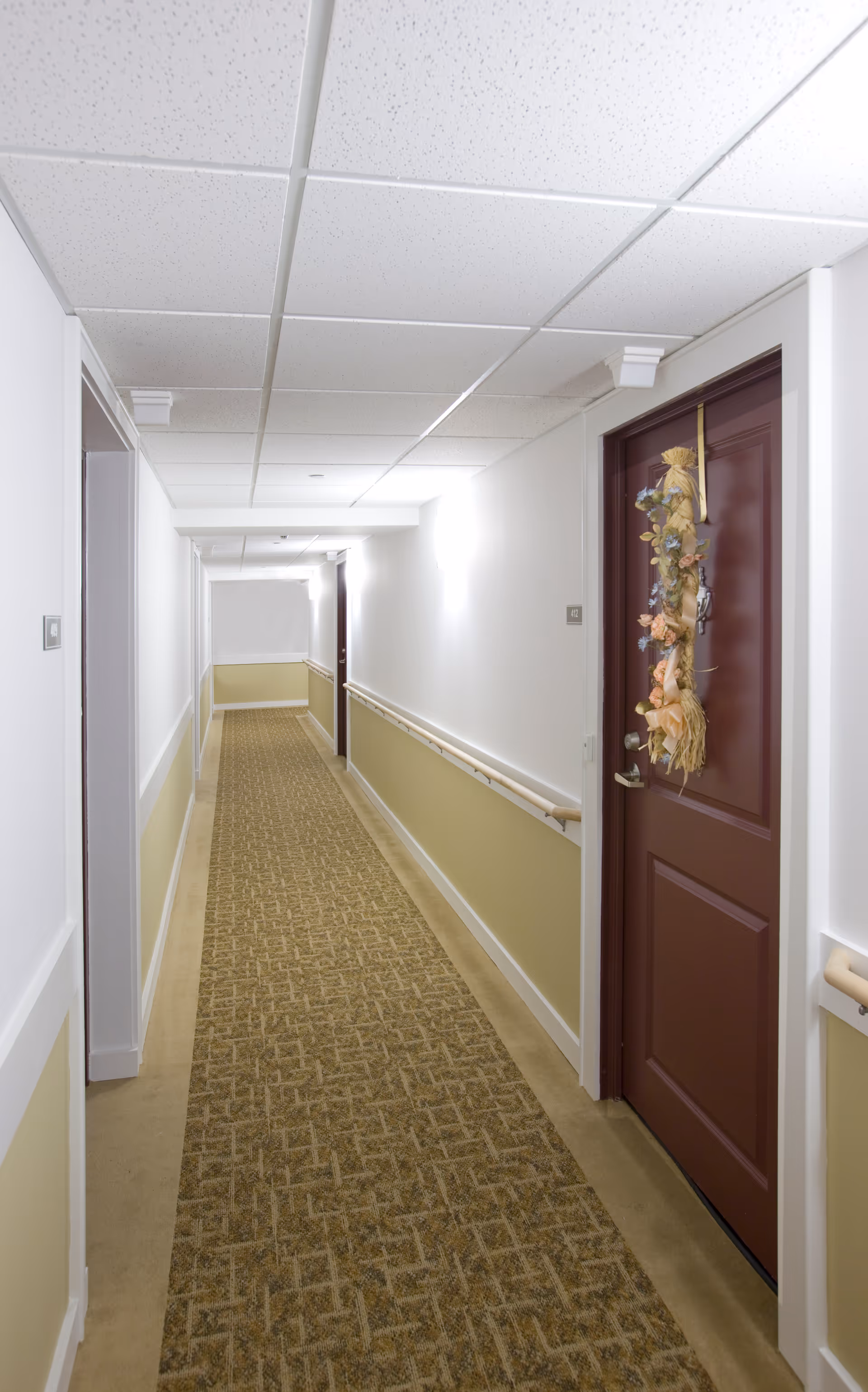 A long, well-lit hallway in a senior living facility with beige and white walls, a patterned carpet, and handrails on both sides. A maroon door on the right side is decorated with a floral wreath.