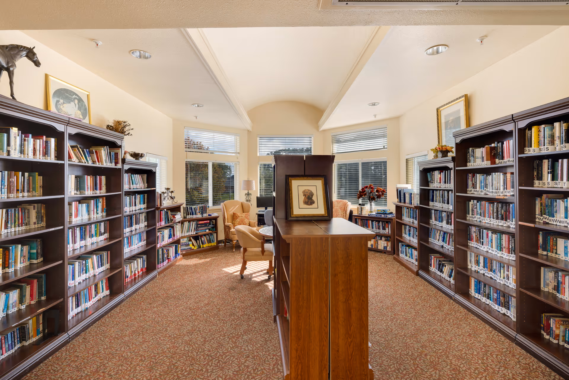 A bright and cozy library room with tall wooden bookshelves filled with books on both sides. The room has large windows letting in natural light, a carpeted floor with a patterned design, and comfortable armchairs near the windows. A framed picture and decorative items are placed on top of the bookshelves.