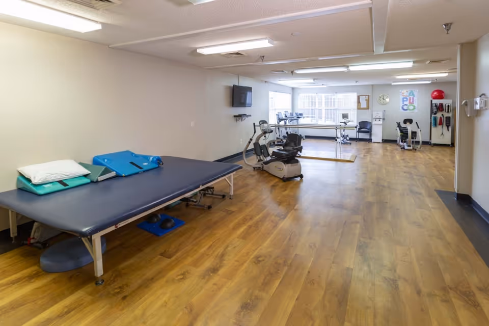 A spacious rehabilitation room with wooden flooring, featuring a padded therapy table with pillows and wedges on the left, exercise equipment including stationary bikes and treadmills in the center and background, a wall-mounted TV, and large windows letting in natural light.
